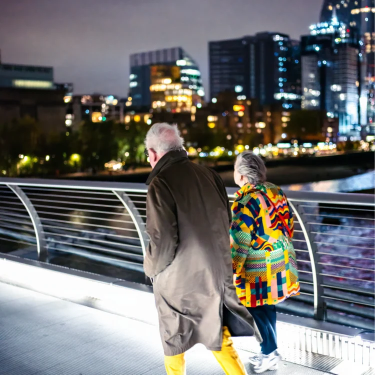 Elderly couple in colourful clothing walking across Millennium Bridge at night – London street photography print